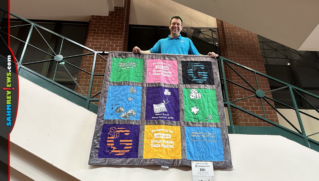 During Great Plains Game Festival, Great Plains Gaming Project president poses with a quilt created by a charity. - SahmReviews.com